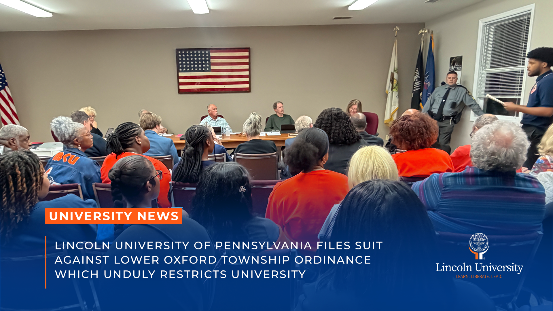 Township board meeting room with officials seated at a table facing a full audience, many wearing orange in a show of Lincoln University community support. A uniformed officer stands near a side door. Lincoln University news graphic overlaid at bottom with headline 'Lincoln University of Pennsylvania Files Suit Against Lower Oxford Township Ordinance Which Unduly Restricts University' and the Lincoln University logo. 1