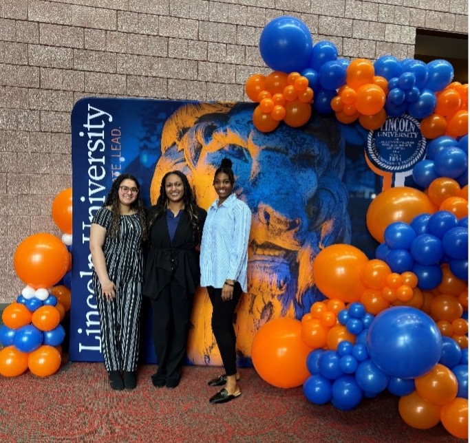 Act 101 director and two students standing in front of a banner surrounded by orange and blue balloons