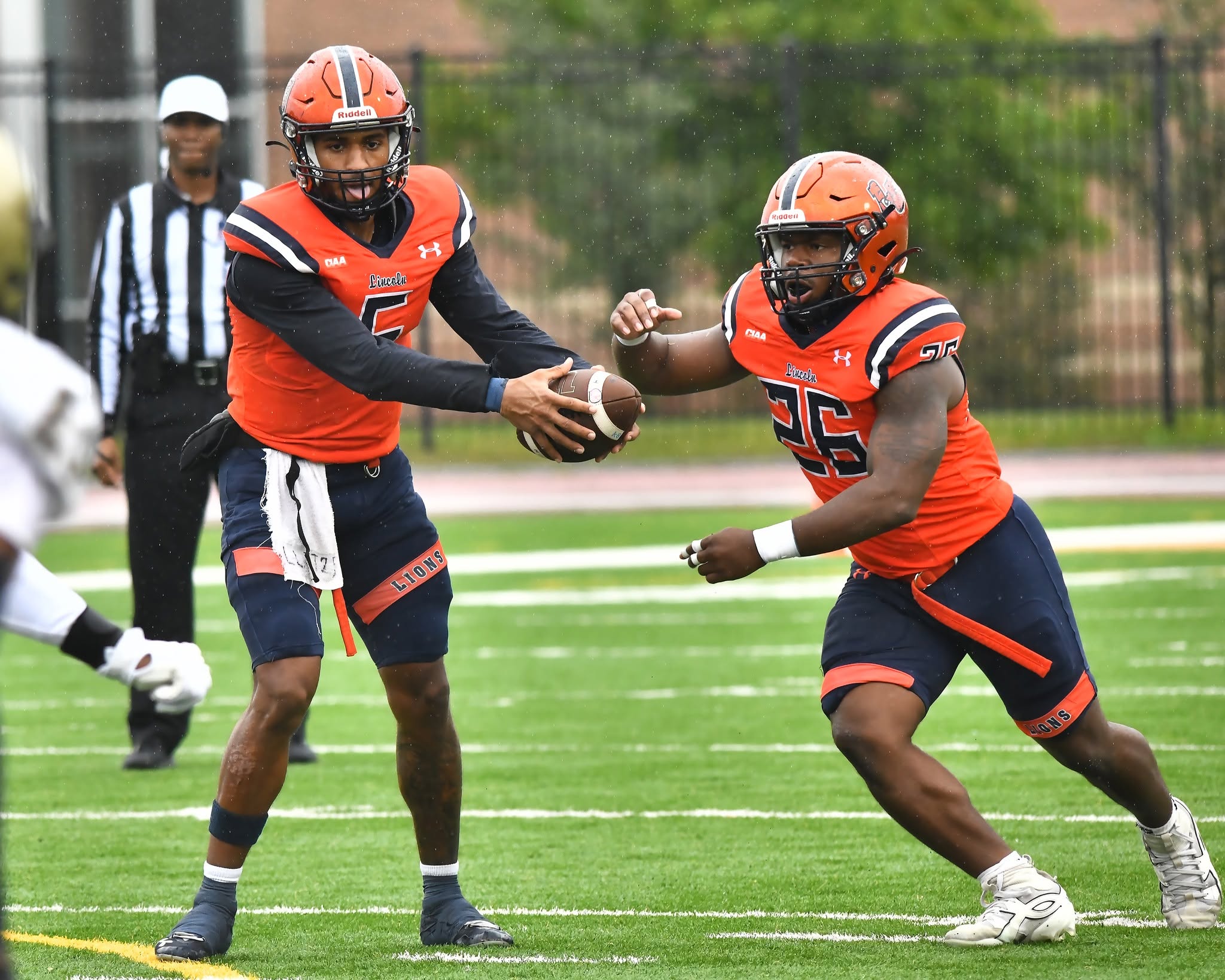 Lincoln University quarterback number 5 hands off to running back number 26 during a rainy football game.