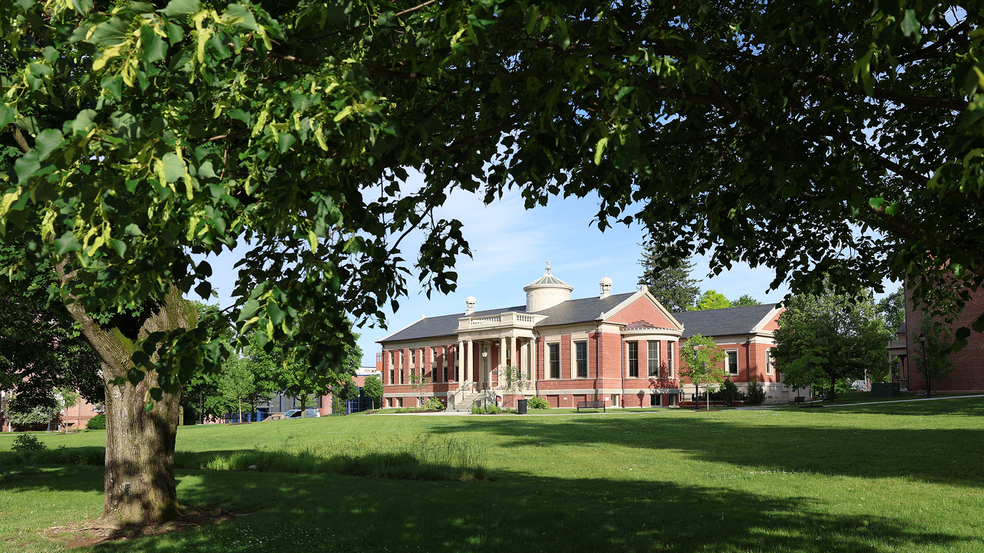 Vail Hall, a red brick building with a white columned entrance and domed cupola, framed by tree branches across a green lawn at Lincoln University.